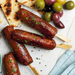 Skewered kofta on a white surface with bread and olives.