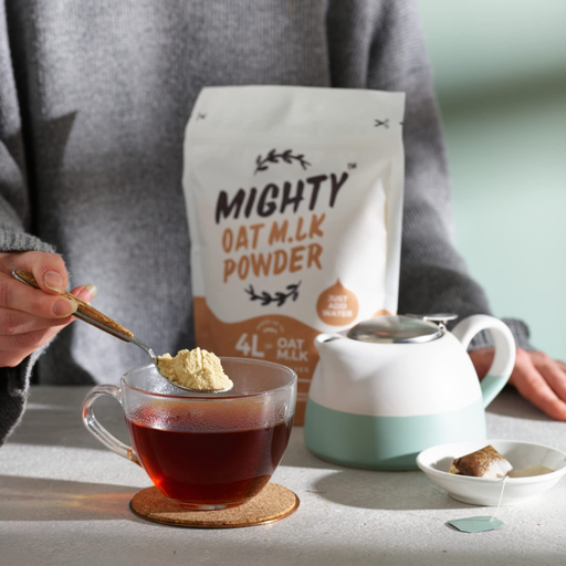 Person adding Mighty Oat Mlk Powder to a cup of tea with a teapot and package in the background.