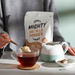 Person adding Mighty Oat Mlk Powder to a cup of tea with a teapot and package in the background.