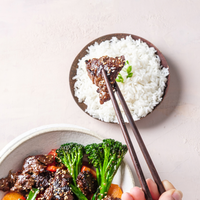 A close-up of a hand holding chopsticks with a piece of glazed vegan mushroom brisket with another plate of white rice and a bowl of stir-fried broccoli and carrots in the background on a pale pink surface.