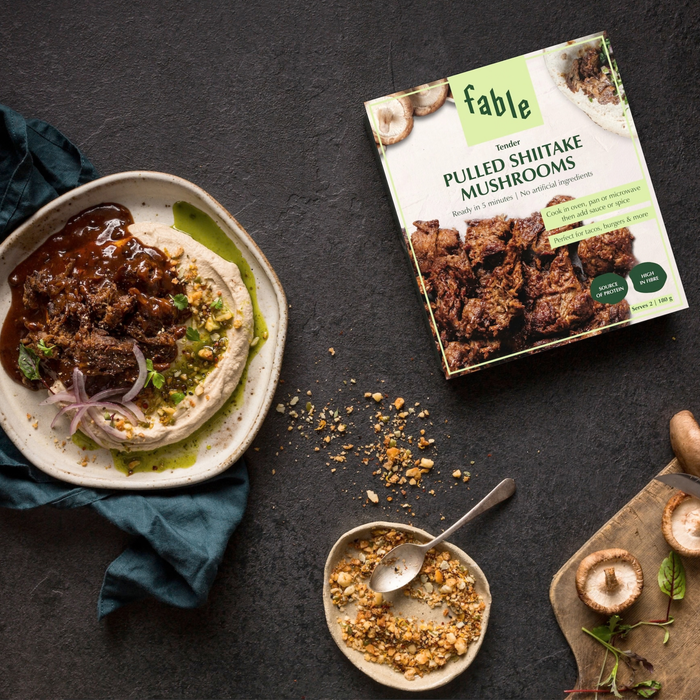A styled food scene on a dark stone surface showing a bowl of hummus topped with pulled shiitake mushrooms and dressing, alongside the Fable brand box of pulled shiitake mushrooms and small bowls of seasoning and mushrooms.