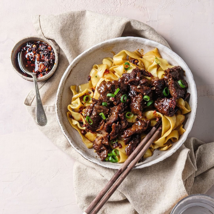 A bowl of shiitake mushrooms and noodle stir-fry with green onions, served on a beige linen napkin with chopsticks and a small side dish of sauce on a beige background.