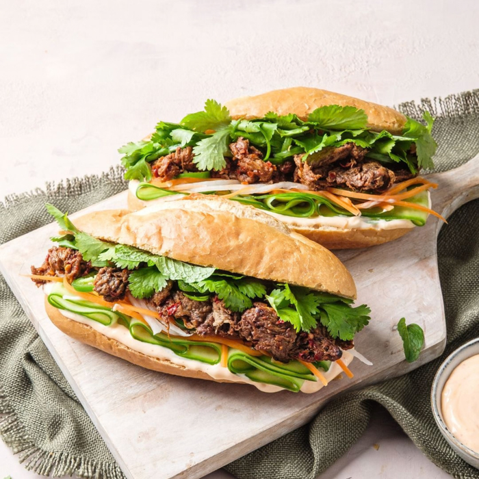 Two large banh mi sandwiches filled with sliced vegan meat, fresh cilantro, shredded carrots, and green onions, served on a wooden board with a small bowl of sauce.