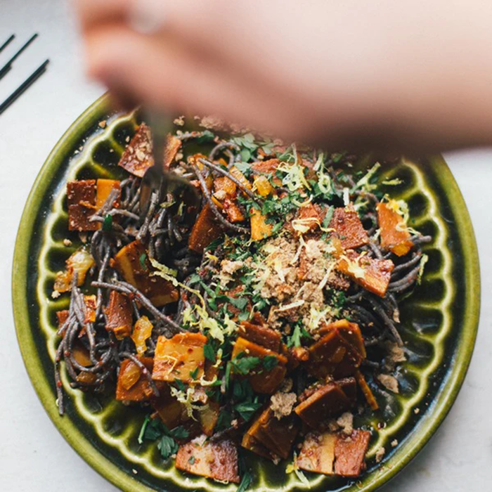 A close-up shot of a hand sprinkling seasoning over a green plate of cooked pasta with vegan bacon rasher, herbs, and cheese on a light background.