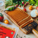 Sliced vegan bacon rashers on a wooden cutting board, surrounded by tomatoes, herbs, bread crust on a wooden table.