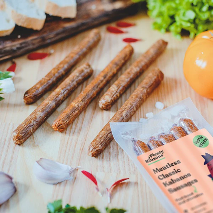 Several vegan kabanos are arranged on a wooden board next to a package of Plenty Delicious Classic Kabazos sausages, with garlic cloves, red petals, and green herbs around them.