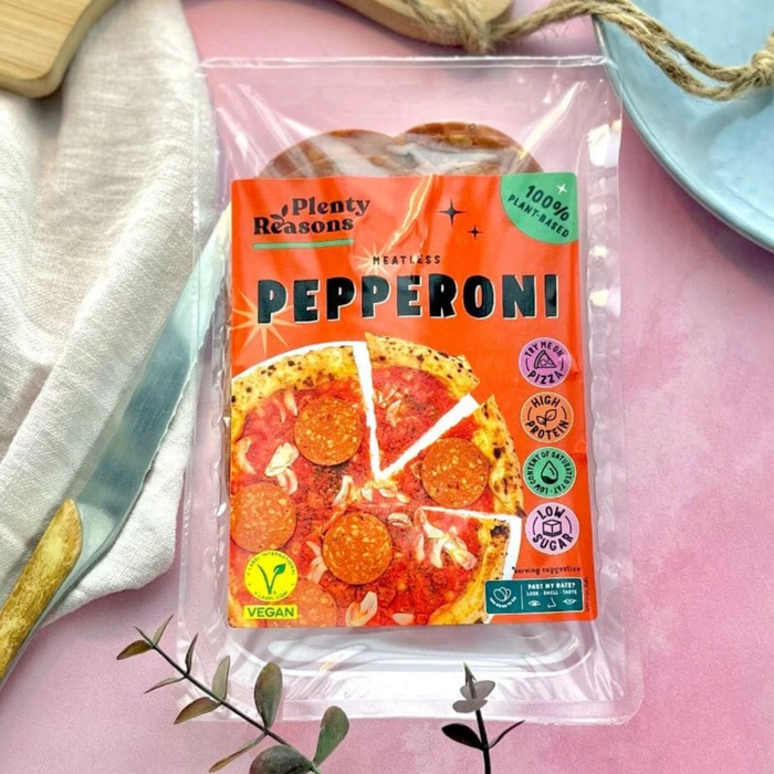 A package of Plenty Reasons Pepperoni vegan slices in a clear plastic clamshell tray, placed on a pink surface with decorative props including a white cloth, a blue plate, and small greenery.