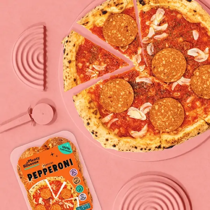 A pink flat-lay featuring a pepperoni pizza on a matching pink plate, with a package of Peppy Peppers pepperoni slices in the lower left and pink pizza cutters around the edges.